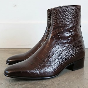May include: A pair of dark brown ankle boots featuring a textured crocodile skin pattern. These boots have a sleek, pointed toe design, a low block heel, and a side zipper closure, showcasing a polished finish with a structured silhouette.