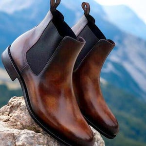May include: A pair of brown leather Chelsea boots with elastic side panels and pull tabs. The boots have a polished finish and are set against a blurred mountain backdrop. The boots are a classic style.