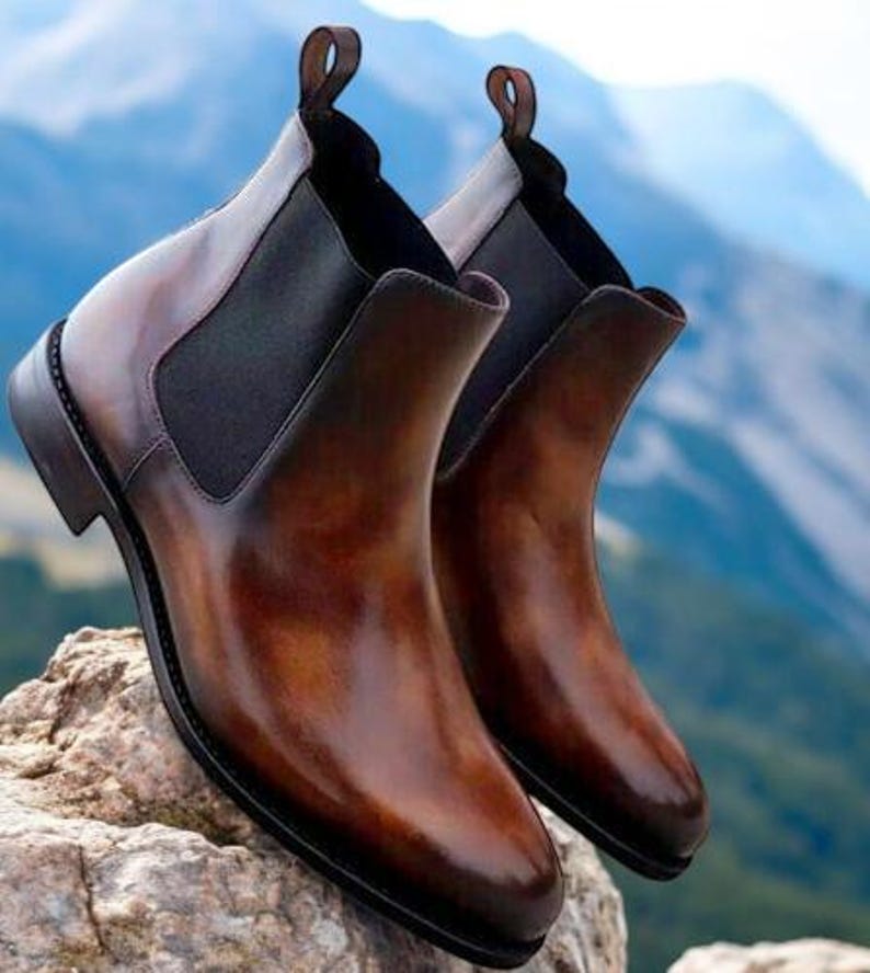 May include: A pair of brown leather Chelsea boots with elastic side panels and pull tabs. The boots have a polished finish and are set against a blurred mountain backdrop. The boots are a classic style.