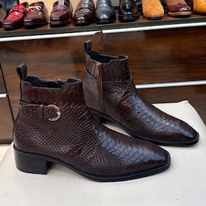Könnte beinhalten: Ein Paar dunkelbraune Stiefeletten mit einer strukturierten Schlangenleder-Optik, einem seitlichen Reißverschluss, einem kleinen Blockabsatz und einem dekorativen Riemen über dem Knöchel, der mit einer goldfarbenen Schnalle an der Außenseite befestigt ist.