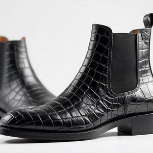 May include: A pair of black Chelsea boots with a crocodile-textured pattern. The boots have a pull tab at the back and a low heel. The boots are made of a shiny, leather-like material.