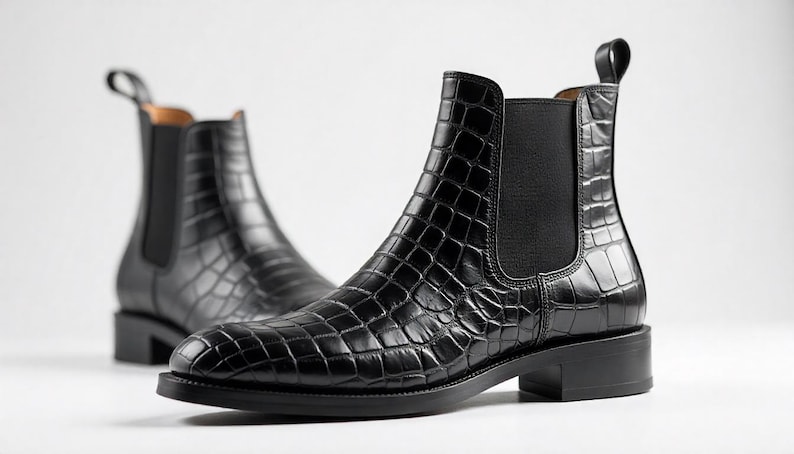 May include: A pair of black Chelsea boots with a crocodile-textured pattern. The boots have a pull tab at the back and a low heel. The boots are made of a shiny, leather-like material.