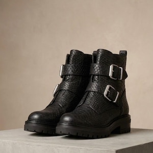 Puede incluir: Un par de botines negros con un patrón texturizado de piel de serpiente, punteras redondeadas y suelas gruesas con tacos. Cada bota está diseñada con dos correas ajustables en la parte delantera, aseguradas con hebillas de metal plateado.