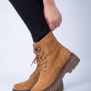 Könnte beinhalten: Ein beigefarbener Schnürstiefel aus Wildleder mit einer dicken, braunen Profilsohle aus Gummi. Der Stiefel verfügt über Metallösen, passende beige Schnürsenkel und ein kleines silbernes sternförmiges Emblem an der Außenseite nahe der Ferse.
