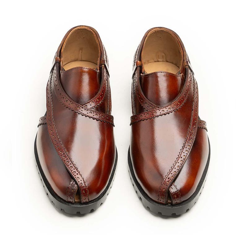 May include: A pair of brown leather shoes with a polished appearance. The shoes have a distinctive design with decorative stitching and a crisscross strap. The soles are black, and the shoes are displayed against a white backdrop.