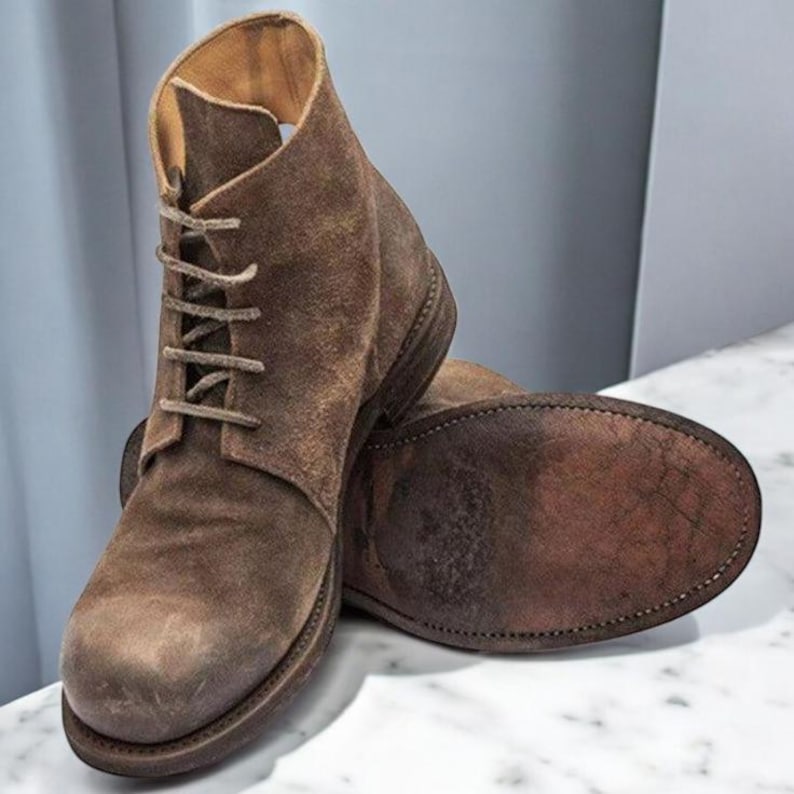 May include: Brown suede ankle boots with a lace-up design. The boots have a worn, textured appearance and a sturdy sole. The boots are displayed on a marble surface.