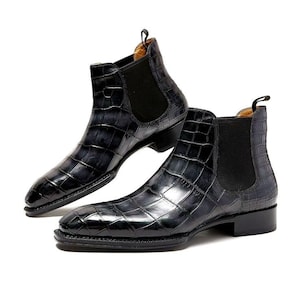 May include: A pair of black Chelsea boots with a crocodile skin pattern. The boots have a pointed toe, elastic side panels, and a small pull tab at the back. The boots are made of leather.