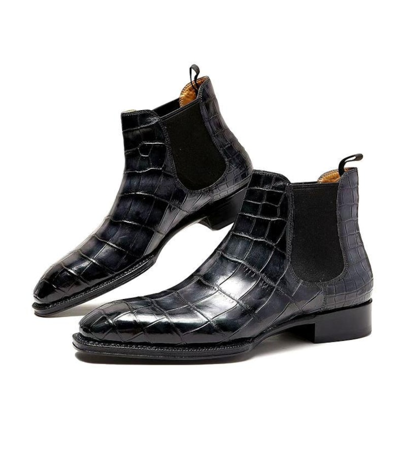 May include: A pair of black Chelsea boots with a crocodile skin pattern. The boots have a pointed toe, elastic side panels, and a small pull tab at the back. The boots are made of leather.