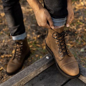 May include: Brown leather boots with black laces and metal eyelets. The boots have a cap toe design and are worn with dark denim jeans. The boots are on a wooden surface, with a blurred background of leaves.