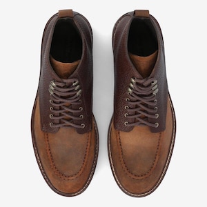 May include: A pair of brown leather shoes with a two-tone design. The upper part is a darker brown, textured leather, while the lower part is a lighter brown suede. The shoes have brown laces and a classic, casual style.