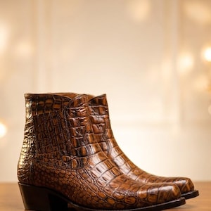 Puede incluir: Un par de botines marrones con un patrón texturizado de piel de cocodrilo. Las botas tienen punta afilada, un tacón de bloque apilado y un forro interior de cuero liso. La superficie exterior presenta un acabado brillante con tonos marrón oscuro.