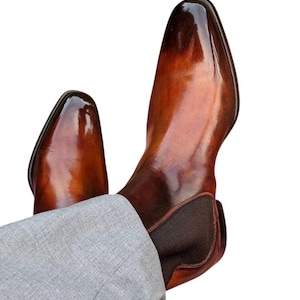 May include: A pair of brown leather Chelsea boots with a glossy finish. The boots have a pointed toe and elastic side panels. The boots are worn with gray trousers.