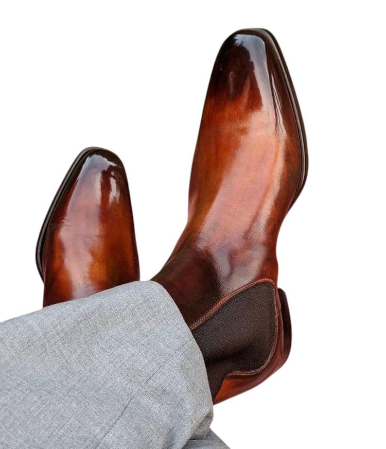 May include: A pair of brown leather Chelsea boots with a glossy finish. The boots have a pointed toe and elastic side panels. The boots are worn with gray trousers.
