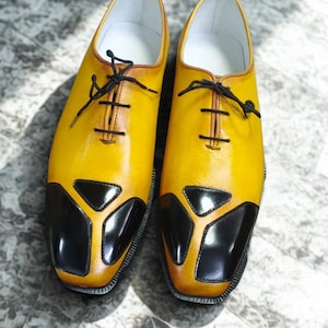Puede incluir: Un par de zapatos de vestir de cuero amarillo con detalles negros en la puntera y un diseño clásico con cordones. Los zapatos tienen un acabado pulido y un interior claro.