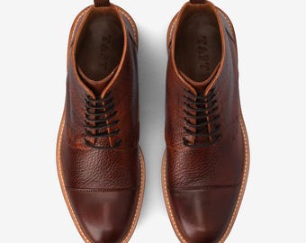 May include: A pair of brown leather boots with a textured finish, black laces, and a rounded toe. The boots have a light-colored sole and the word "TAFT" is visible inside the boot.