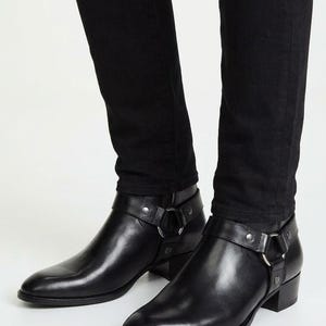 May include: Black leather ankle boots featuring a pointed toe, a low block heel, and a harness strap detail across the instep. The strap is secured with silver-toned metal hardware, including a circular ring and rivets, worn with black denim jeans.