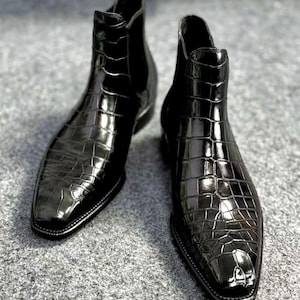 May include: A pair of black ankle boots featuring a glossy finish and a textured crocodile skin pattern. These boots have a pointed toe, a high shaft, and a sleek silhouette with a visible seam running down the centre of the front vamp.
