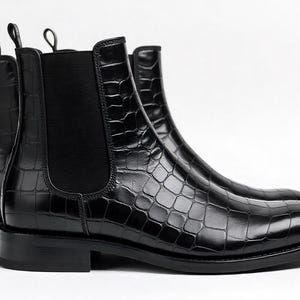May include: A pair of black Chelsea boots with a crocodile-textured pattern. The boots have a sleek, polished appearance, with elastic side panels and pull tabs. The boots are set against a white background.
