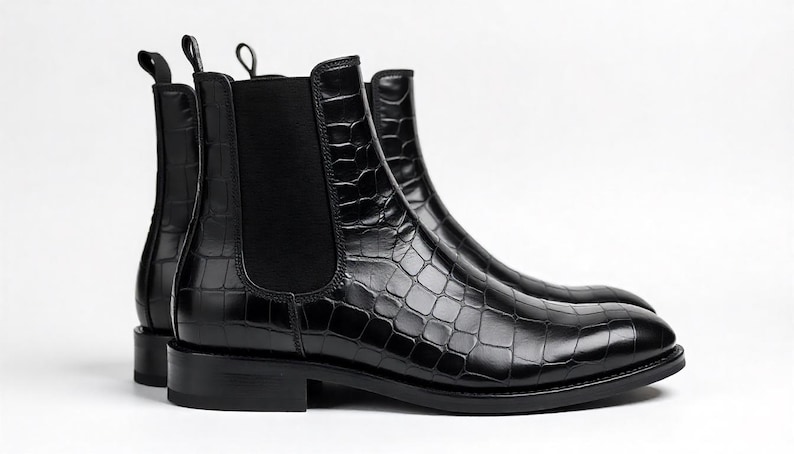 May include: A pair of black Chelsea boots with a crocodile-textured pattern. The boots have a sleek, polished appearance, with elastic side panels and pull tabs. The boots are set against a white background.