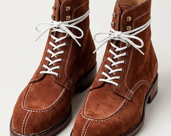 May include: A pair of brown suede boots featuring a moc-toe construction with prominent white contrast stitching, white laces, and brass-toned metal eyelets. The boots have a stacked leather heel and a dark brown sole with a visible welt.