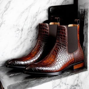 Könnte beinhalten: Ein Paar braune Chelsea-Stiefel aus Leder mit Krokodilleder-Textur. Die Stiefel haben elastische Seiteneinsätze und Zuglaschen. Die Stiefel werden auf einer Marmoroberfläche präsentiert.