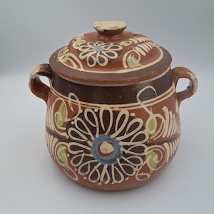 Puede incluir: Una olla de cerámica redonda con tapa y dos asas. La olla es principalmente marrón con diseños florales blancos, azules y verdes. La tapa también presenta motivos florales. La olla tiene un aspecto rústico y artesanal.