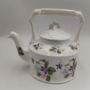 May include: A white ceramic teapot featuring a floral pattern of purple, blue, and pink violets with green leaves. The teapot has a sculpted handle, a curved spout, and a lid topped with a molded floral finial, all in a glossy white finish.