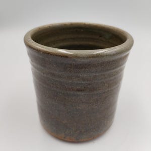 May include: A cylindrical ceramic cup featuring a mottled brown and grey glaze with visible horizontal ridges along the exterior surface. The rim is smooth and rounded, transitioning into a slightly darker, earthy tone at the base of the vessel.