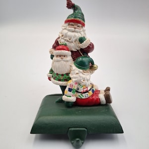 May include: A festive Christmas stocking holder with three Santa Claus figurines. The Santas wear red, green, and white outfits, complete with white beards and hats. The holder is green and has a hook for hanging.