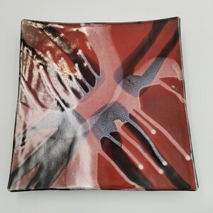 May include: A square glass plate featuring a glossy finish with an abstract design in shades of deep red, black, and white. The surface displays overlapping brushstroke-like patterns that create a layered effect across the entire square dish.