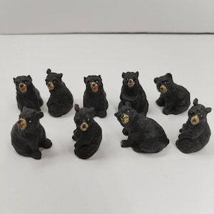 May include: A collection of eight small, black bear figurines in various seated poses. Each bear is crafted with a textured finish, featuring painted tan details on the snout and inner ears, with some bears showing open mouths and others with closed mouths.
