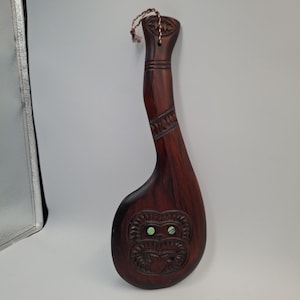 May include: A dark brown wooden club with a long handle and a wide, rounded head. The handle features carved geometric patterns and a braided cord at the top. The head displays a carved face with two inlaid iridescent green shell eyes.