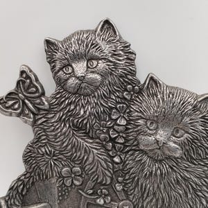 Può includere: Una spilla in metallo color argento raffigurante due gattini dal pelo folto posizionati fianco a fianco. Una farfalla è appoggiata vicino all'orecchio del gattino a sinistra. Entrambi i gattini sono circondati da piccoli dettagli floreali.