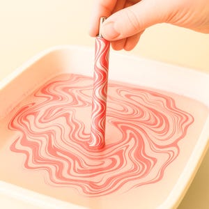 May include: A red and white patterned pen is held above a tray of pink liquid, creating a swirling pattern. The pen has a silver top and a repeating wave design. The liquid in the tray is also swirled with the same red and white pattern.