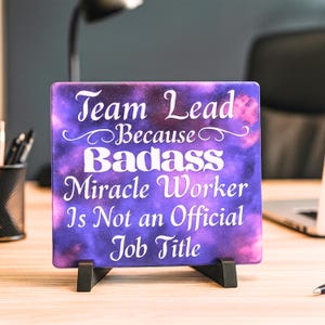 Puede incluir: Un letrero decorativo con el texto "Team Lead Because Badass Miracle Worker Is Not an Official Job Title" en letras blancas sobre un fondo de galaxia púrpura y rosa. El letrero se muestra en un soporte negro.