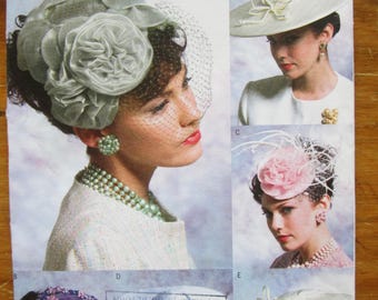 May include: A Vogue Accessories pattern showcasing vintage hat designs. The image displays a selection of hats, including a gray floral headpiece with a veil, a white hat with a bow, and a pink floral hat. The pattern is labelled V8052.