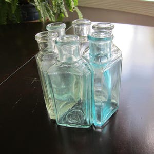 May include: A cluster of six clear, light blue-tinted glass bottles arranged in a hexagonal formation. Each bottle features a rectangular body with rounded corners, a short neck, and a circular opening at the top, creating a unified glass structure.