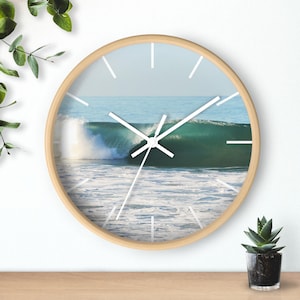 May include: A round wall clock with a light wood frame. The clock face features a photograph of a breaking ocean wave in shades of teal and white. White clock hands and hour markers complete the design.