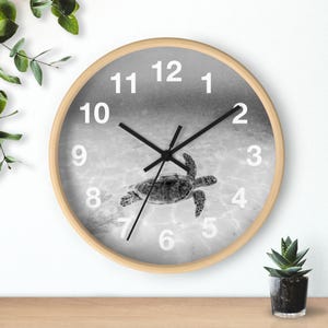 Könnte beinhalten: Runde Wanduhr mit hellem Holzrahmen. Das Zifferblatt zeigt ein Graustufenbild einer Meeresschildkröte, die im Ozean schwimmt. Schwarze Zeiger und weiße Ziffern markieren die Zeit.