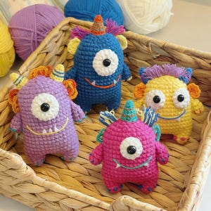 Può includere: Quattro giocattoli a forma di mostro all'uncinetto colorati in un cestino di vimini. I mostri sono blu, viola, gialli e rosa, ognuno con un occhio grande, una bocca sorridente e un piccolo corno. Sullo sfondo sono visibili gomitoli di lana.