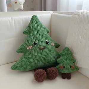 May include: A green crocheted Christmas tree pillow with a smiling face, blush, and black eyes. A smaller matching tree and two brown crocheted balls are also present. The items are on a white surface.