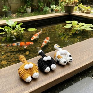 Puede incluir: Tres adornos de culos de gato de ganchillo en amarillo, negro y blanco con traseros blancos y ojos saltones. Los adornos se exhiben sobre una superficie de madera con un estanque de carpas koi en el fondo.