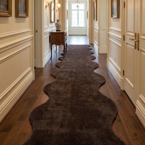 May include: A dark brown, organically shaped rug stretches along a hallway with wooden flooring. The walls are painted cream and decorated with framed pictures. A decorative table and chandelier are visible in the distance.
