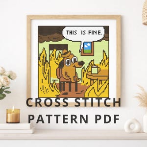 May include: A square cross stitch pattern featuring a pixelated dog sitting at a table in a room filled with yellow flames. The dog has a speech bubble above its head containing the text THIS IS FINE. The design is framed in a light wood frame.