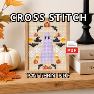 May include: A cross stitch pattern PDF featuring a central light purple ghost surrounded by orange pumpkins, black bats, witch hats, candy corn, and white spider webs, all framed in a light wood frame with the text CROSS STITCH PATTERN PDF.