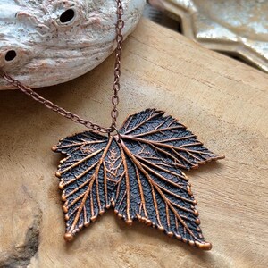 Op de afbeelding: Een ketting met een donkere, getextureerde bladvormige hanger met koperkleurige nerven en randen, hangend aan een bijpassende koperkleurige metalen ketting. De hanger vertoont ingewikkelde, verhoogde botanische details op het oppervlak.