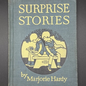 Puede incluir: Libro antiguo titulado "SURPRISE STORIES" de Marjorie Hardy. La portada es de color verde azulado con letras doradas y una ilustración circular de dos niños. Uno lee un libro y el otro señala.