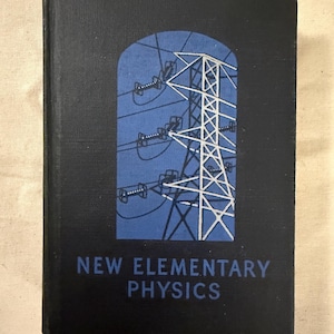 Op de afbeelding: Een zwart hardcover boek met de titel "NEW ELEMENTARY PHYSICS". De omslag toont een blauwe boogvormige illustratie van een hoogspanningsmast en elektrische draden. De tekst is wit.