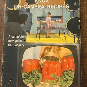 以下が含まれることがあります： 「KNOX ON-CAMERA RECIPES」と題されたヴィンテージの料理本の表紙。スタジオキッチンで料理をする女性のレトロなイラストが描かれています。表紙には、赤いゼラチン型とテキスト「A completely new guide to Gel-Cookery」が表示されたテレビ画面が描かれています。