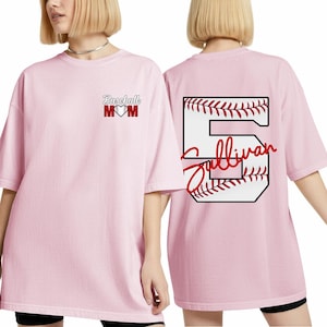 Könnte beinhalten: Hellrosa Baseball-T-Shirt. Auf der Vorderseite steht "Baseball MOM" mit einem Herz. Auf der Rückseite ist eine Baseball-Nummer 5 mit dem Namen "Sullivan" in roter Schrift abgebildet.
